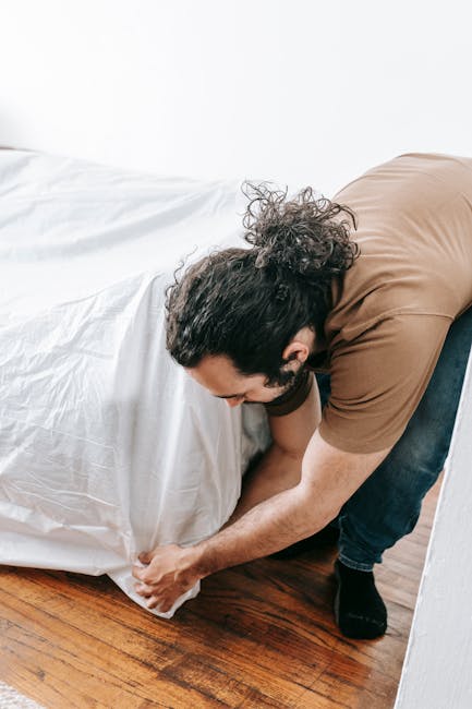 A man with dark, curly hair and a beard, wearing a brown T-shirt and dark jeans, is crouched down on a wooden floor, grasping the edge of a large white plastic bag or tarp. The bag appears to be fille