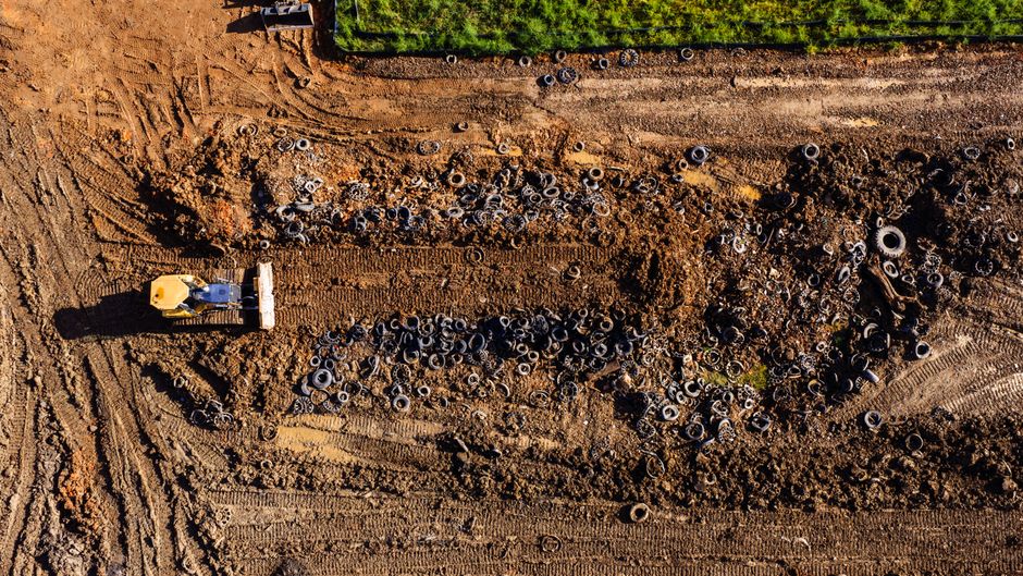 An aerial view of a construction site showing a large expanse of uneven, brown earth with visible tire tracks and soil compression marks. Scattered across the area are numerous discarded black rubber 